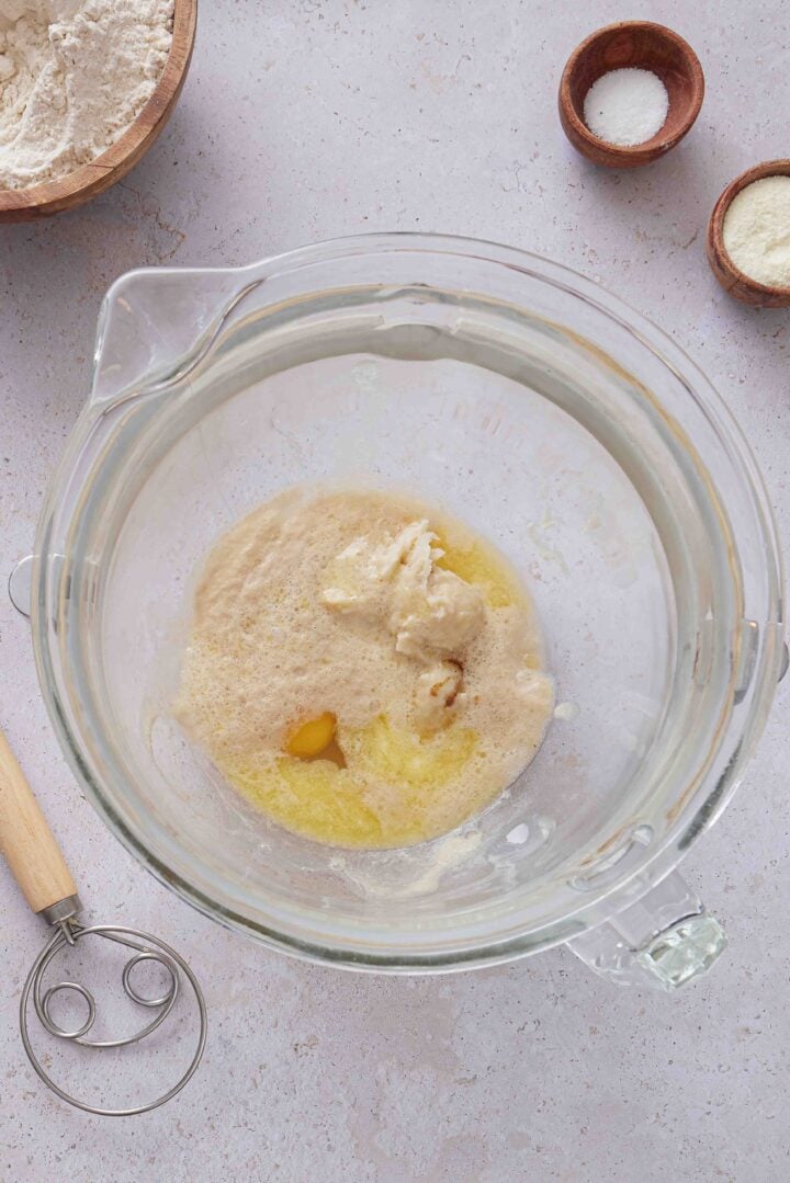 Overhead photo of the wet ingredients for Brazilian sweet condensed milk buns in a glass mixing bowl, including yeast mixture, egg, melted butter, vanilla, and tangzhong.