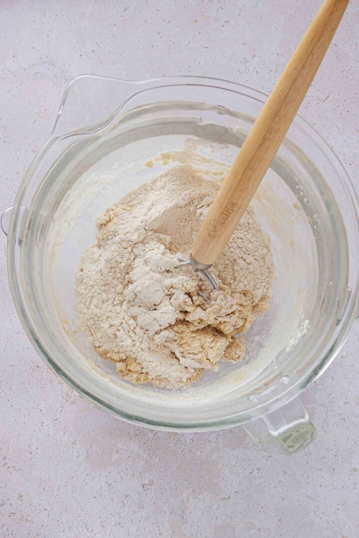Overhead photo of flour being mixed into the wet ingredients for Brazilian sweet condensed milk buns in a glass bowl.