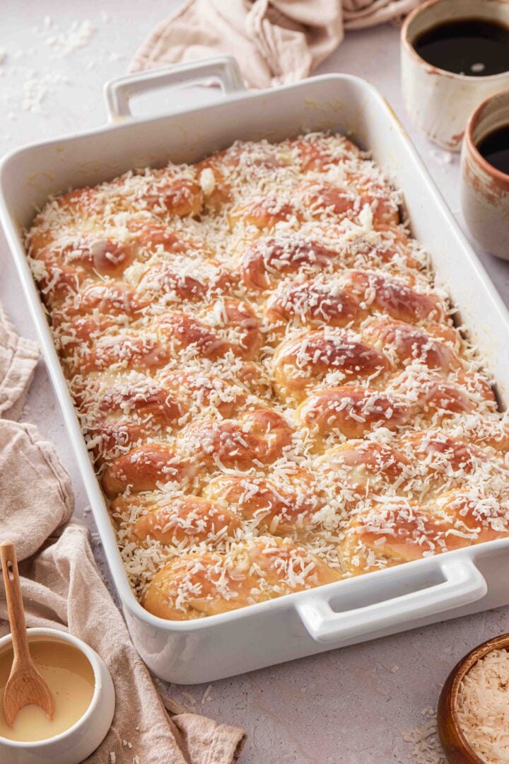 Overhead photo of Brazilian sweet condensed milk buns in a white baking dish, topped with shredded coconut and sweetened condensed milk glaze, with coffee and extra coconut on the side.
