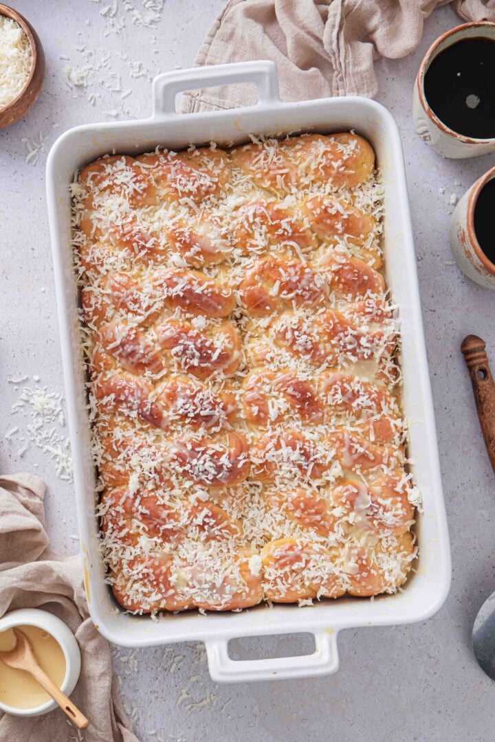 Overhead photo of Brazilian sweet condensed milk buns baked in a white baking dish, glazed with sweetened condensed milk and topped with shredded coconut, with cups of black coffee on the side.