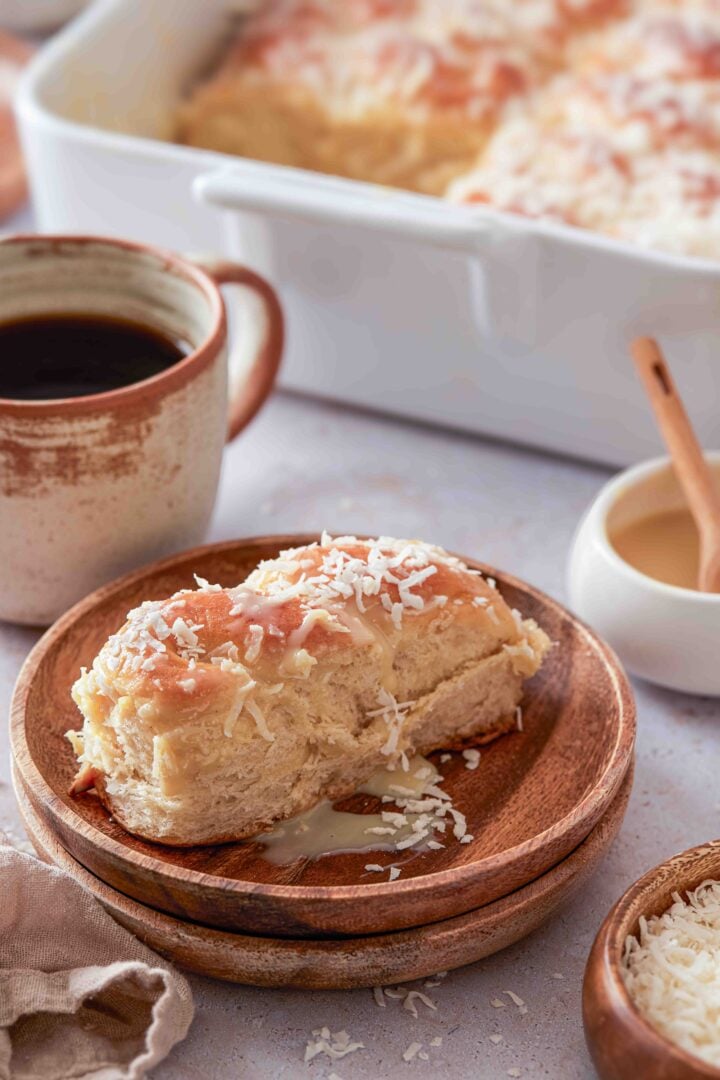 lose-up of a Brazilian sweet condensed milk bun on a wooden plate, topped with shredded coconut and sweetened condensed milk glaze, served with black coffee.