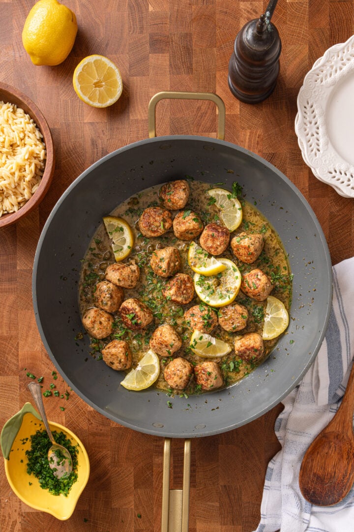 Chicken piccata meatballs in skillet with a bowl of orzo and lemon wedges on wooden table