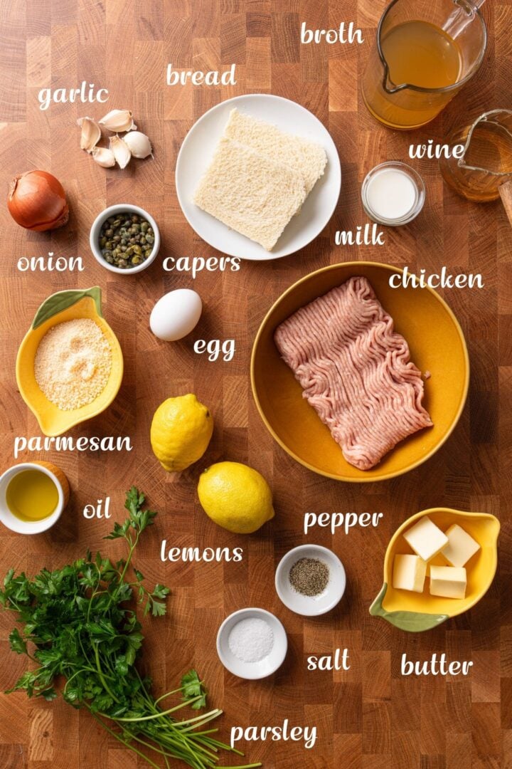 Ingredients for chicken piccata meatballs arranged on a wooden surface, including ground chicken, bread, garlic, onion, capers, egg, Parmesan, lemons, parsley, butter, broth, white wine, milk, olive oil, salt, and pepper