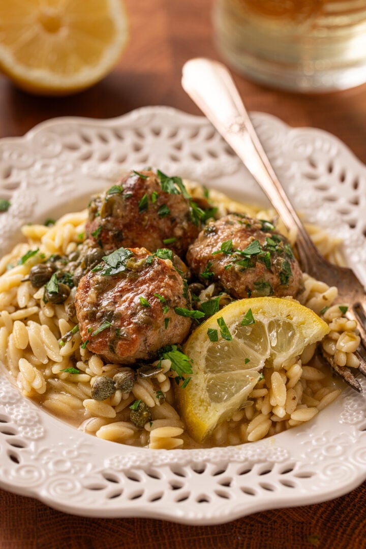Chicken piccata meatballs served over orzo pasta with lemon wedge, capers, and parsley on a white plate