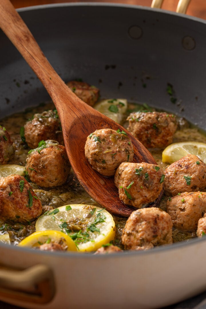Close-up of chicken piccata meatballs in skillet with wooden spoon lifting a meatball from lemon caper sauce