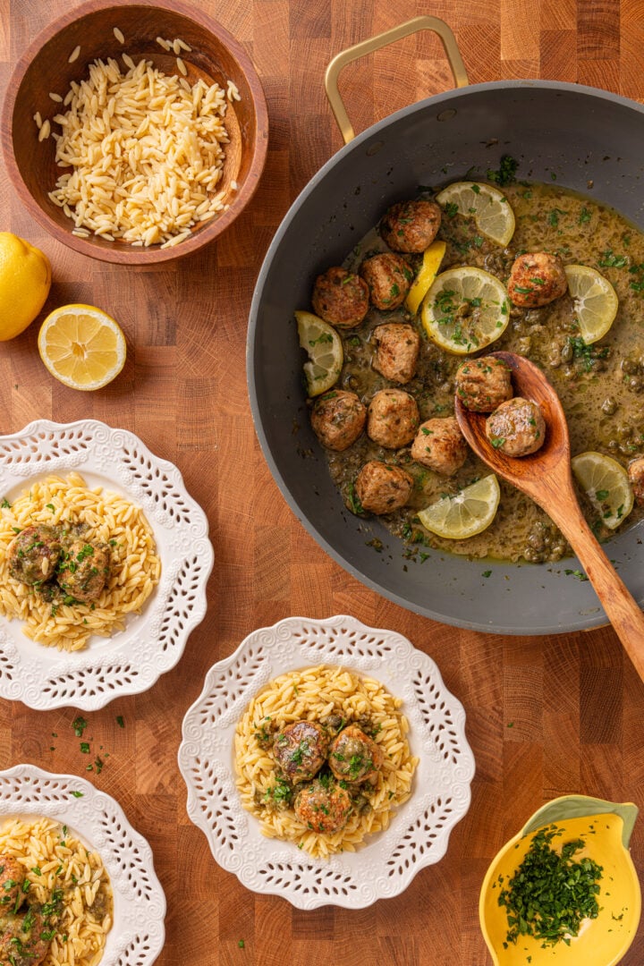 Overhead table with multiple plates of chicken piccata meatballs served over orzo alongside skillet and fresh ingredients