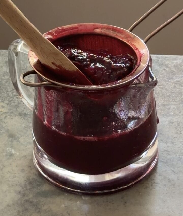 Thick berry sauce being strained into a glass pitcher for a smoother texture.