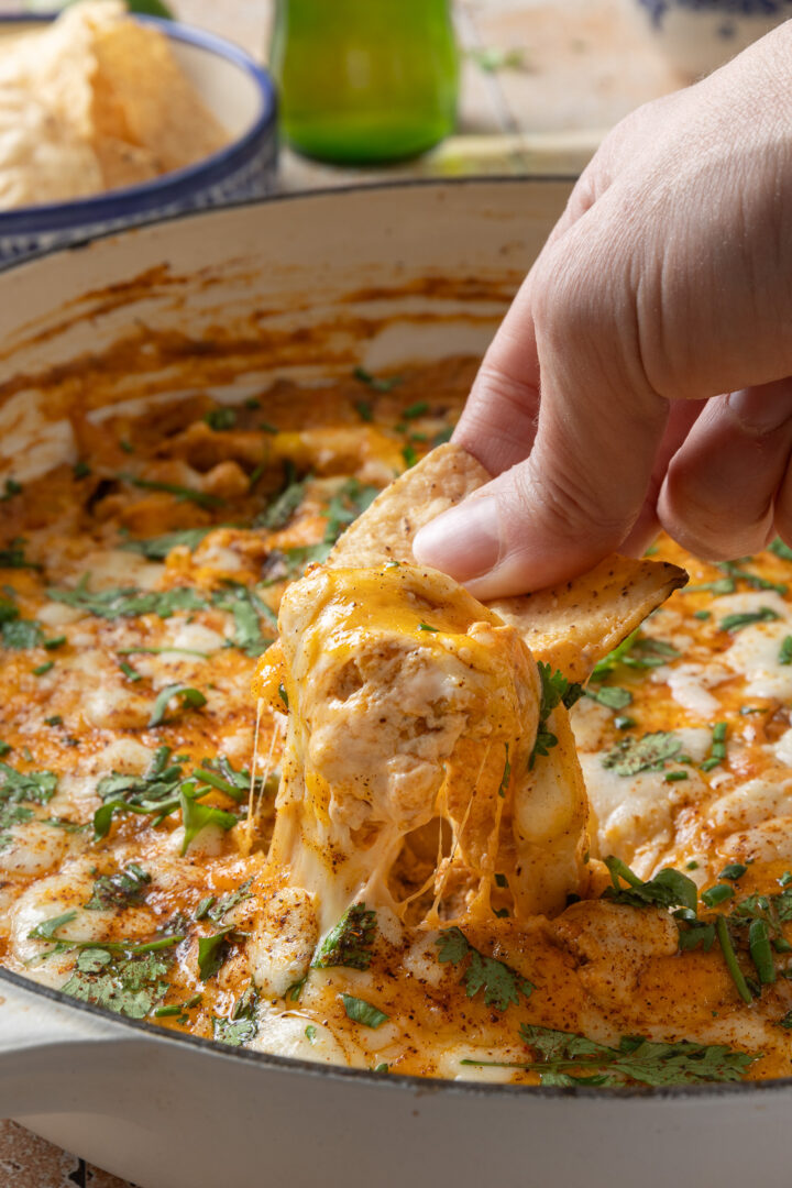 Hand dipping a tortilla chip into creamy Mexican street corn dip with melted cheese and corn in a skillet