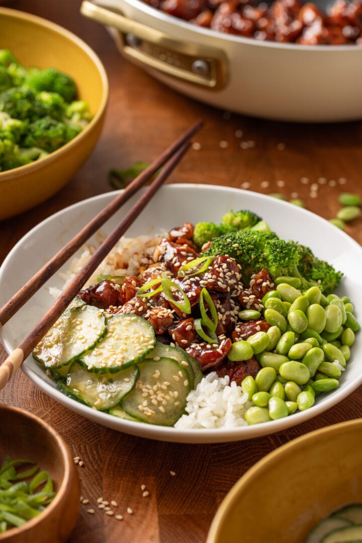Chicken teriyaki rice bowl with rice, broccoli, edamame, and cucumber salad topped with sesame seeds and green onions.