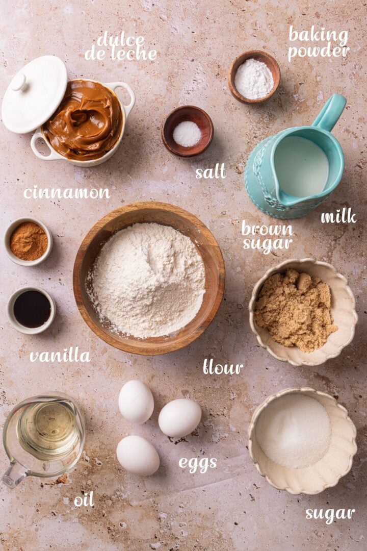 Ingredients for churro cake including flour, eggs, sugar, brown sugar, cinnamon, milk, oil, vanilla, baking powder, salt, and dulce de leche arranged on a countertop.