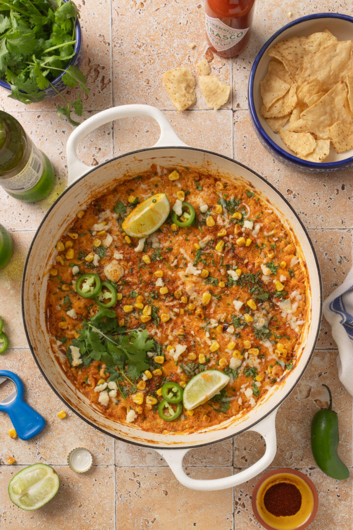 Overhead view of baked Mexican street corn dip in a skillet with tortilla chips, lime wedges, and cilantro