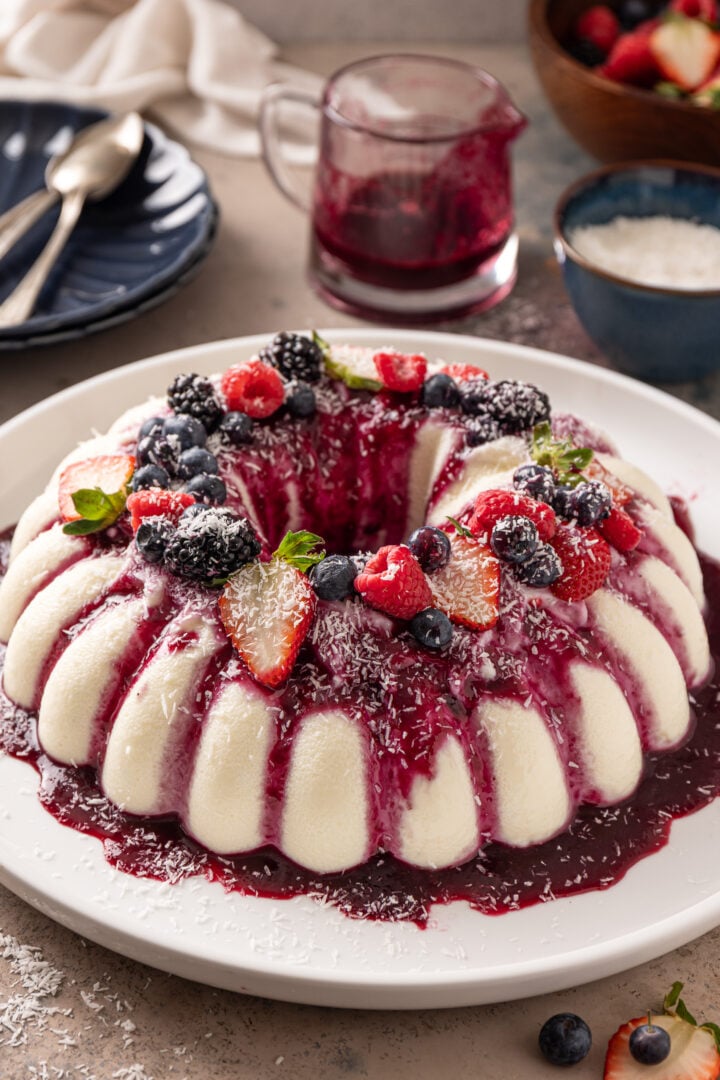 Whole coconut flan with berry sauce, fresh berries, and shredded coconut served on a white platter.