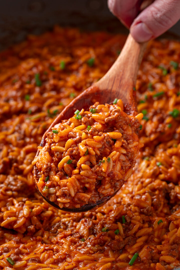 Spoon lifting creamy ground beef orzo with tomato cream sauce and cheddar cheese from a skillet.