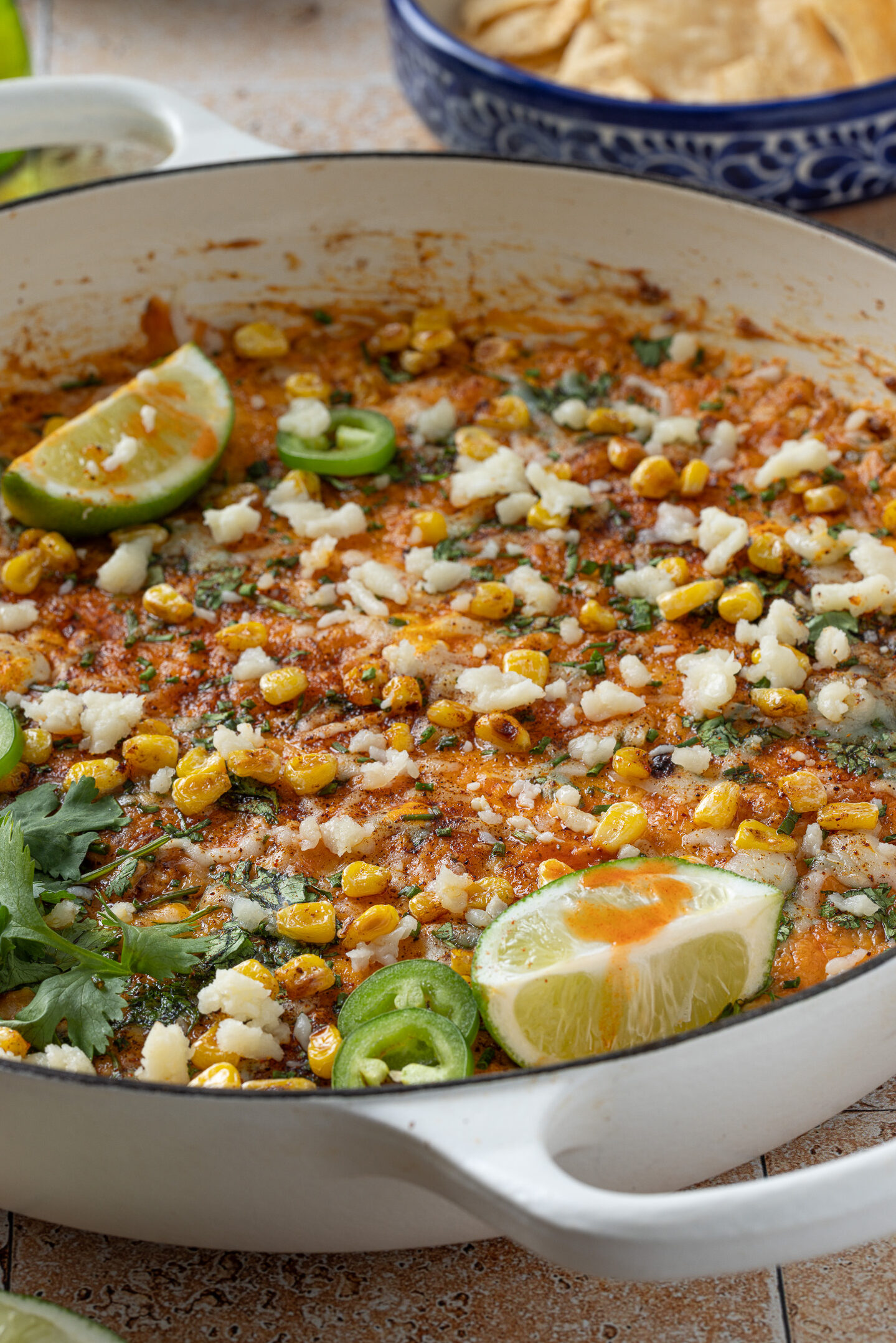 Baked Mexican street corn dip topped with cotija cheese, cilantro, jalapeños, and lime wedges in a skillet