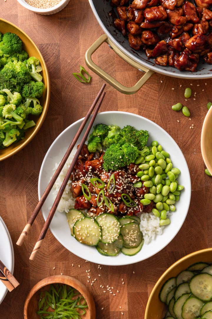 Chicken teriyaki rice bowl topped with sesame seeds and green onions, served with broccoli, edamame, and cucumber salad.