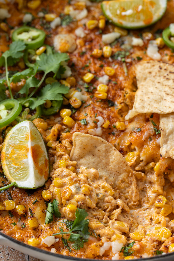 Close-up of tortilla chip scooping creamy corn dip with melted cheese and jalapeños