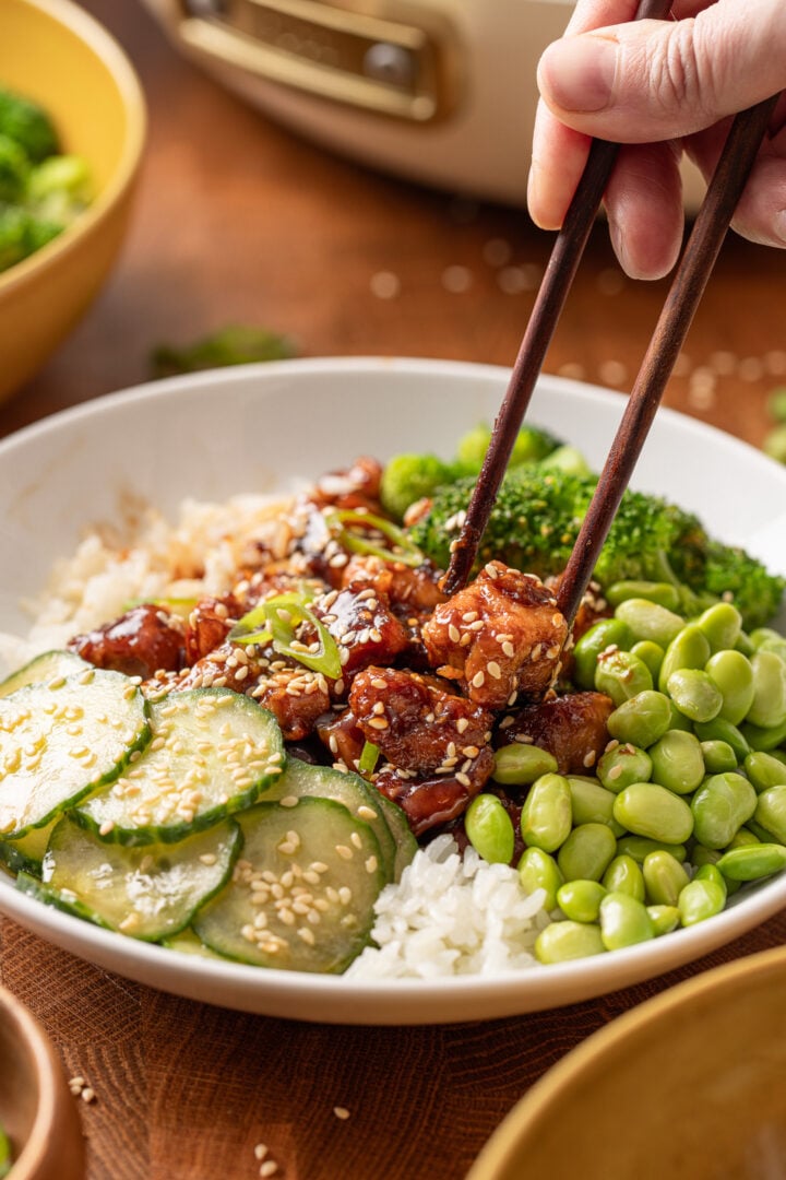 Chopsticks lifting glossy homemade teriyaki chicken from a rice bowl with broccoli, rice, and cucumber salad.