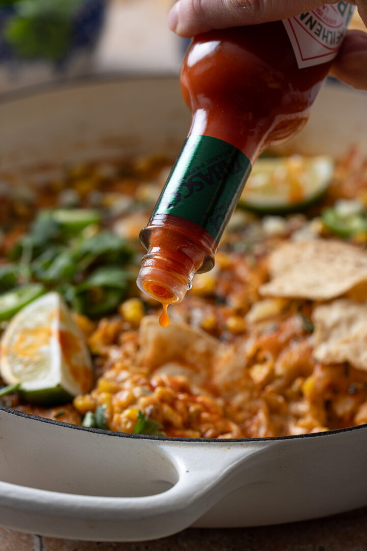 Hot sauce being drizzled over creamy Mexican street corn dip in a skillet