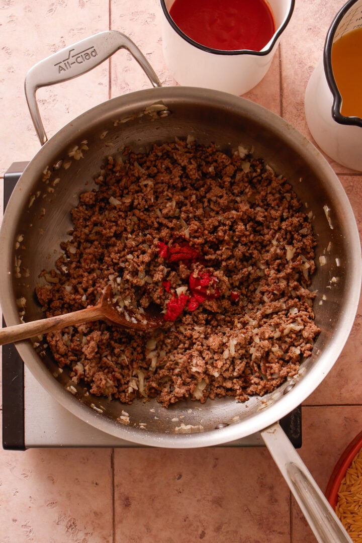 Tomato paste being added to browned ground beef and onions in a skillet.