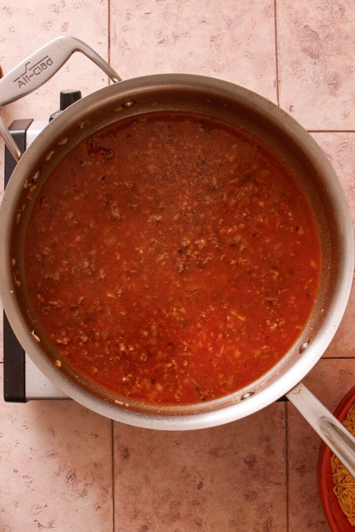 Tomato and beef broth mixture simmering in a skillet for creamy ground beef orzo.