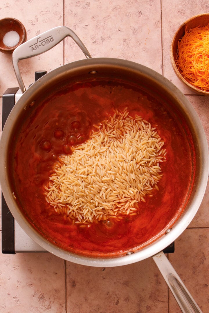 Dry orzo being added to tomato broth mixture for one-pot creamy ground beef orzo.