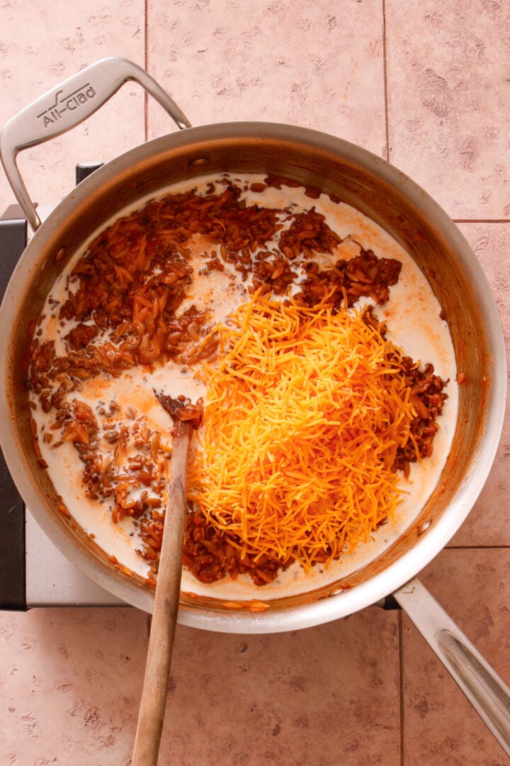 Heavy cream and shredded cheddar cheese being added to creamy tomato ground beef orzo.