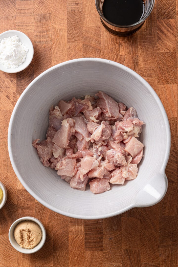 Raw chicken pieces in a bowl with ingredients for homemade teriyaki chicken on a wooden surface.
