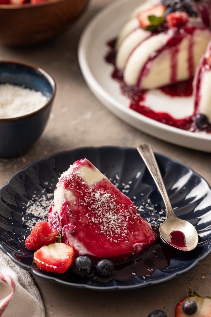 Slice of Brazilian coconut flan with berry sauce, shredded coconut, and fresh berries served on a dark blue dessert plate.