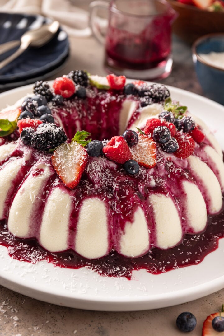 Brazilian coconut flan (manjar branco) topped with berry sauce, fresh berries, and shredded coconut on a white serving platter.