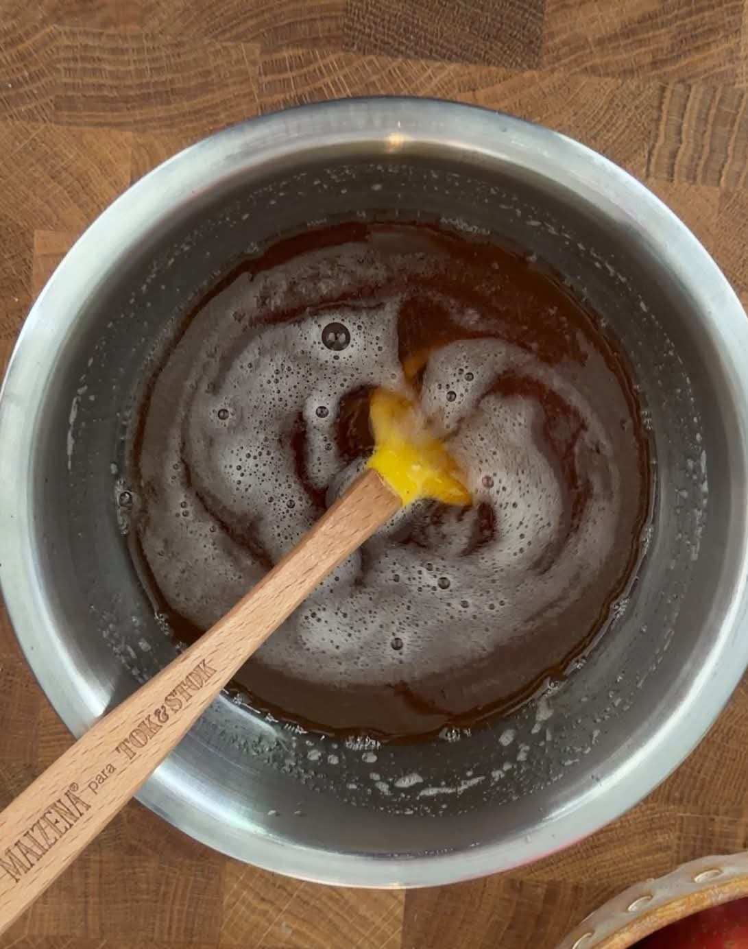 Browned butter in a metal bowl with a spatula, surrounded by fresh apples and vanilla on a wooden work surface.