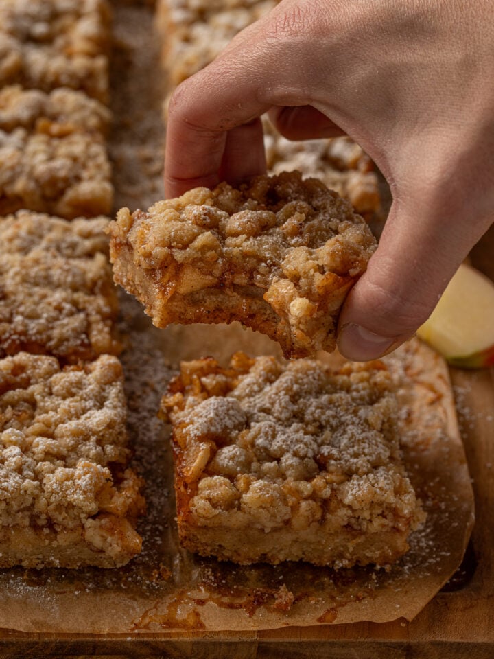 Hand holding an apple crumble bar showing layers of brown butter crust, spiced apple filling and crumble topping.