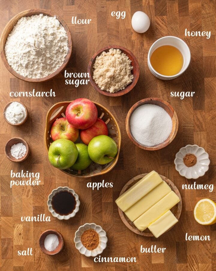 Ingredients for apple crumble bars arranged on a wooden surface, including apples, flour, butter, sugars, honey, egg, lemon and spices.