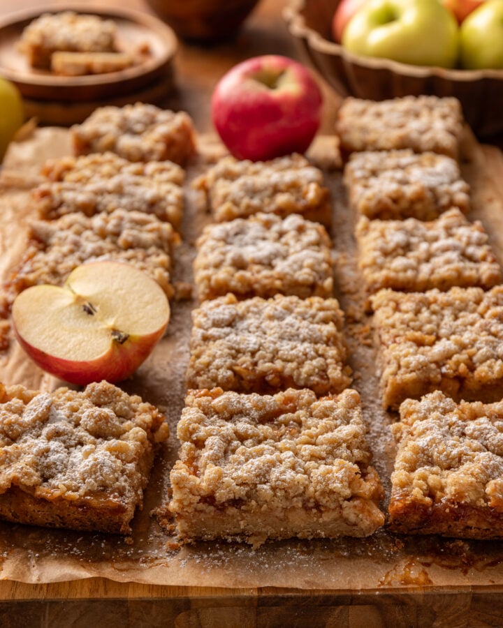 Homemade apple crumble bars arranged on parchment with fresh apples and a dusting of powdered sugar.