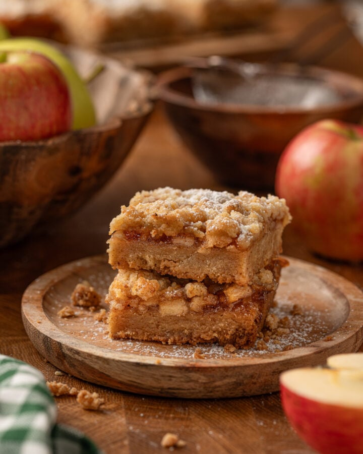 Two apple crumble bars stacked on a wooden plate with fresh apples and powdered sugar.