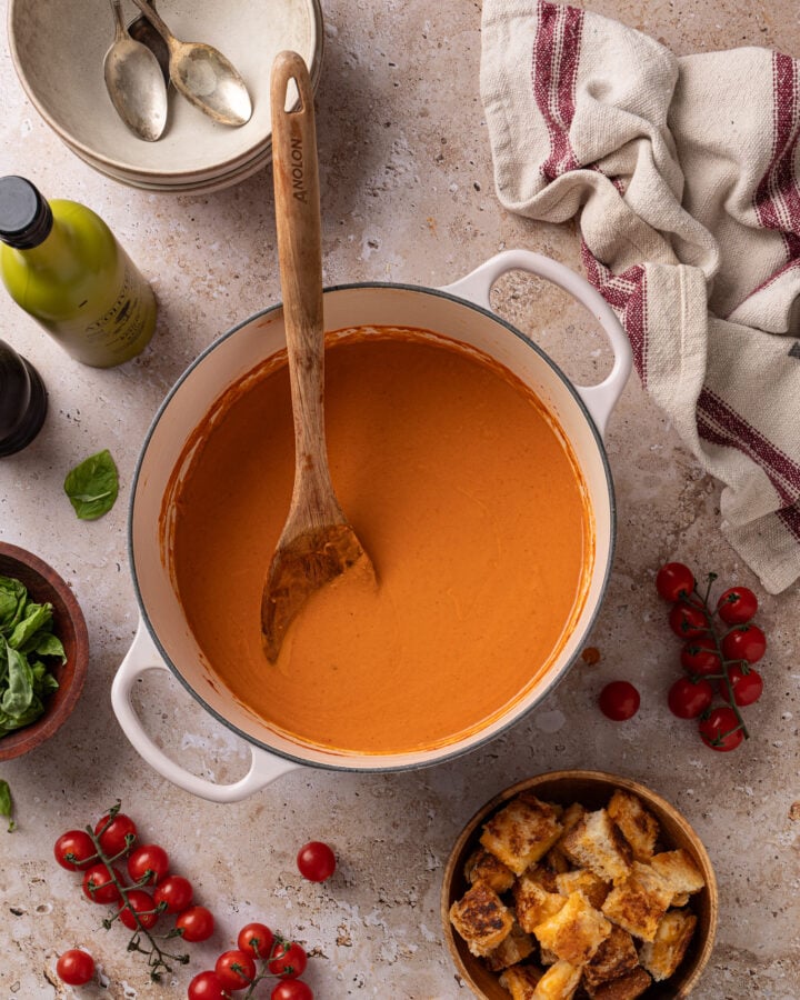 Overhead view of creamy roasted tomato soup in a white Dutch oven with a wooden spoon, with grilled cheese croutons and fresh tomatoes alongside.