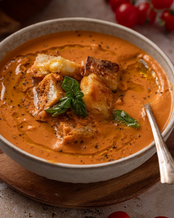 Close-up of roasted tomato soup topped with golden grilled cheese croutons, fresh basil, cracked black pepper, and a drizzle of oil.