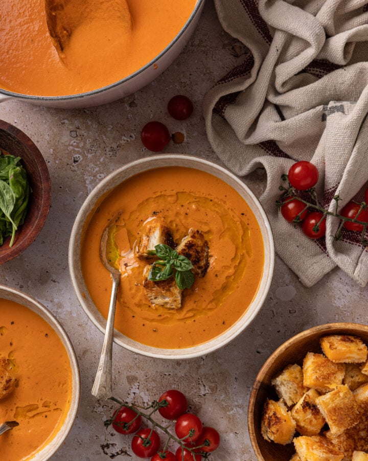 Overhead view of tomato soup garnished with grilled cheese croutons, basil, and oil, with a pot of soup and extra croutons nearby.