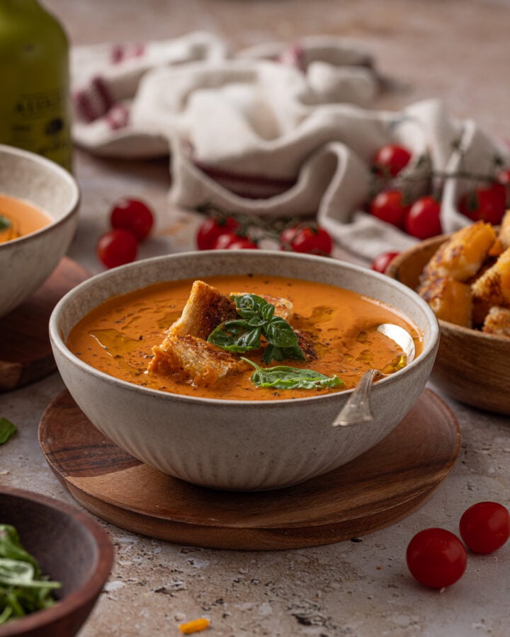 Creamy roasted tomato soup in a gray bowl with grilled cheese croutons, basil, oil, and black pepper, with extra croutons on the side.