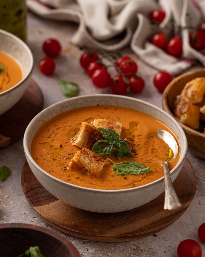 Bowl of roasted tomato soup on a wooden plate, topped with toasted grilled cheese cubes, fresh basil, black pepper, and oil.